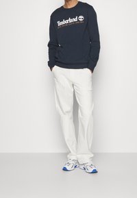 Laivastonsininen puuvillainen collegepaita, jossa "Timberland"-logo valkoisella tekstillä, yhdistetty valkoisiin housuihin ja harmaisiin lenkkareihin, joissa on sinisiä yksityiskohtia.