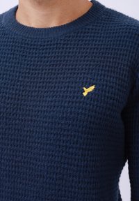 Pull en maille bleu marine avec un motif texturé et un col rond. Présente un petit emblème d'oiseau jaune sur la poitrine gauche.