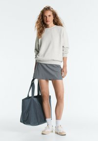 Femme aux cheveux ondulés portant un sweat-shirt gris clair, une jupe courte rayée, des chaussettes blanches, des baskets, tenant un grand sac cabas sombre, debout sur fond blanc.