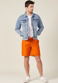 Giacca di jeans con lavaggio blu, t-shirt bianca e pantaloni corti arancioni. I pantaloni corti hanno un cordino e sono lunghi fino al ginocchio. Sneakers bianche completano l'outfit.