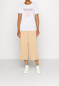 Hvid bomulds t-shirt med en pink og sort trekantet GUESS-logo; parret med beige plisserede culotter og hvide sneakers.