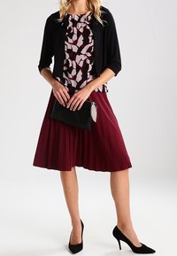 Black cardigan over a patterned blouse in shades of pink, paired with a pleated burgundy skirt and a black clutch with a silver accent.