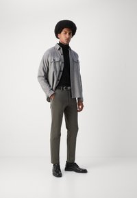 Chemise grise à boutons sur un col roulé noir, pantalon vert olive et chaussures noires. Le modèle se tient devant un fond blanc.