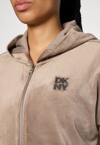 Beige krūšu jakas ar mīkstu, samtainu tekstūru. Tam ir kapuce un mirdzoša logo uzlīme ar rhinestones kreisajā pusē.