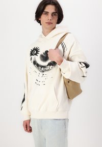 Kremfarget hettegenser med svarte abstrakte grafikker og lommer foran. Modellen holder en beige veske. Lyseblå jeans fullfører antrekket.