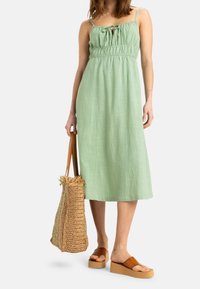 Robe en lin vert clair avec des bretelles spaghetti ajustables, un corsage froncé et un ourlet au genou. Accompagnée d'un sac en paille tissé et de sandales à plateforme.