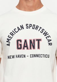 Weißes T-Shirt mit schwarzem und rotem Text "GANT" in der Mitte, umgeben von "American Sportswear" oben und "New Haven - Connecticut" unten.