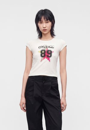 Młoda kobieta o czarnych włosach nosi biały krótki t-shirt z "Obey Studios 89" i wielokolorową grafiką gwiazdy, połączony z czarnymi spodniami.