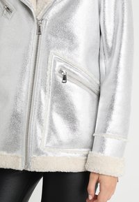 Giacca metallica argento con fodera in shearling visibile sul colletto, sui polsini e sull'orlo, dotata di zip frontale e tasca con zip inclinata sul lato destro.