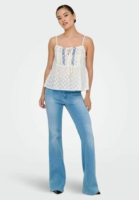 Top peplum blanco con detalles bordados azules y lazo en el frente, combinado con jeans acampanados de color azul claro. Tela suave con un patrón texturizado.