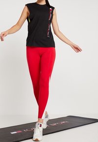 Persona che indossa un top senza maniche nero e leggings rossi, che cammina su un tappetino da esercizio nero con il testo e il logo "Tommy Sport".