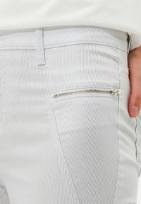 Pantalones de pana en gris claro con sutiles rayas verticales. Presentan un bolsillo con cremallera en el muslo derecho con herrajes plateados.
