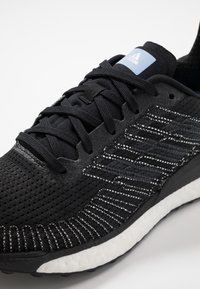Baskets de sport noires avec une tige en mesh texturé et des accents blancs, une semelle rembourrée et un système de laçage traditionnel ; détail du logo bleu.