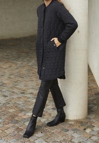 Musta tikattu pitkä takki, jossa painonapit, sivutaskut ja resorit hihansuissa, yhdistetty mustiin nahkahousuihin ja neliönmallisiin nilkkureihin.