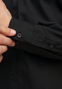 Camicia nera a maniche lunghe con una texture liscia, polsini regolabili con due bottoni neri e un design classico. Il tessuto è ben stirato.
