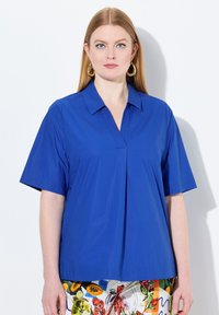 Blusa blu a maniche corte con scollatura a collare e taglio a V. Tessuto morbido, vestibilità rilassata, abbinata a un fondo colorato e fantasia.