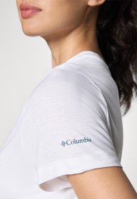 Balta medvilninė marškinėliai su apvalia iškirpte ir trumpomis rankovėmis, kairiajame rankovėje su mėlynu "Columbia" logotipu.