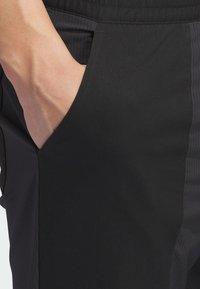 Juodos sportinės kelnės su šonine kišene, lygia medžiagos tekstūra ir vertikaliu juostelių raštu vienoje pusėje.