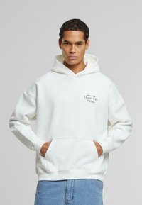Hvid hætte sweatshirt med en frontlomme, løs pasform og et grafisk print med teksten "Middag med Venner" på venstre bryst. Blødt, tekstureret stof.