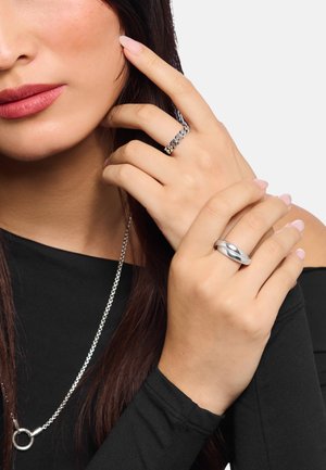 Frau trägt schwarzen schulterfreien Oberteil und silberne Kettenhalskette, zeigt zwei silberne Ringe an ihren Fingern nahe Gesicht und Schulter.