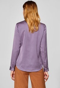 Långärmad, lila blus med diamantmönster och knappade manschetter, med en slät textur och rundad fåll. Bakifrån sedd.