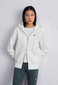 Molete branco com fecho zip, capuz com cordão, bolsos frontais e logo preto da Nike no lado esquerdo do peito. Usado sobre uma t-shirt branca e leggings verdes.