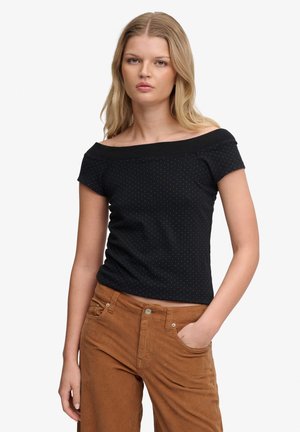 Młoda kobieta o długich blond włosach, ubrana w czarną bluzkę z odkrytymi ramionami w drobne kropki i brązowe spodnie, ręka w kieszeni, neutralny wyraz twarzy.