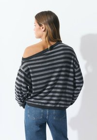 Jersey a rayas en tono gris oscuro y negro, con un corte oversize y un escote amplio. Combinado con jeans azules, mostrando un estilo relajado.