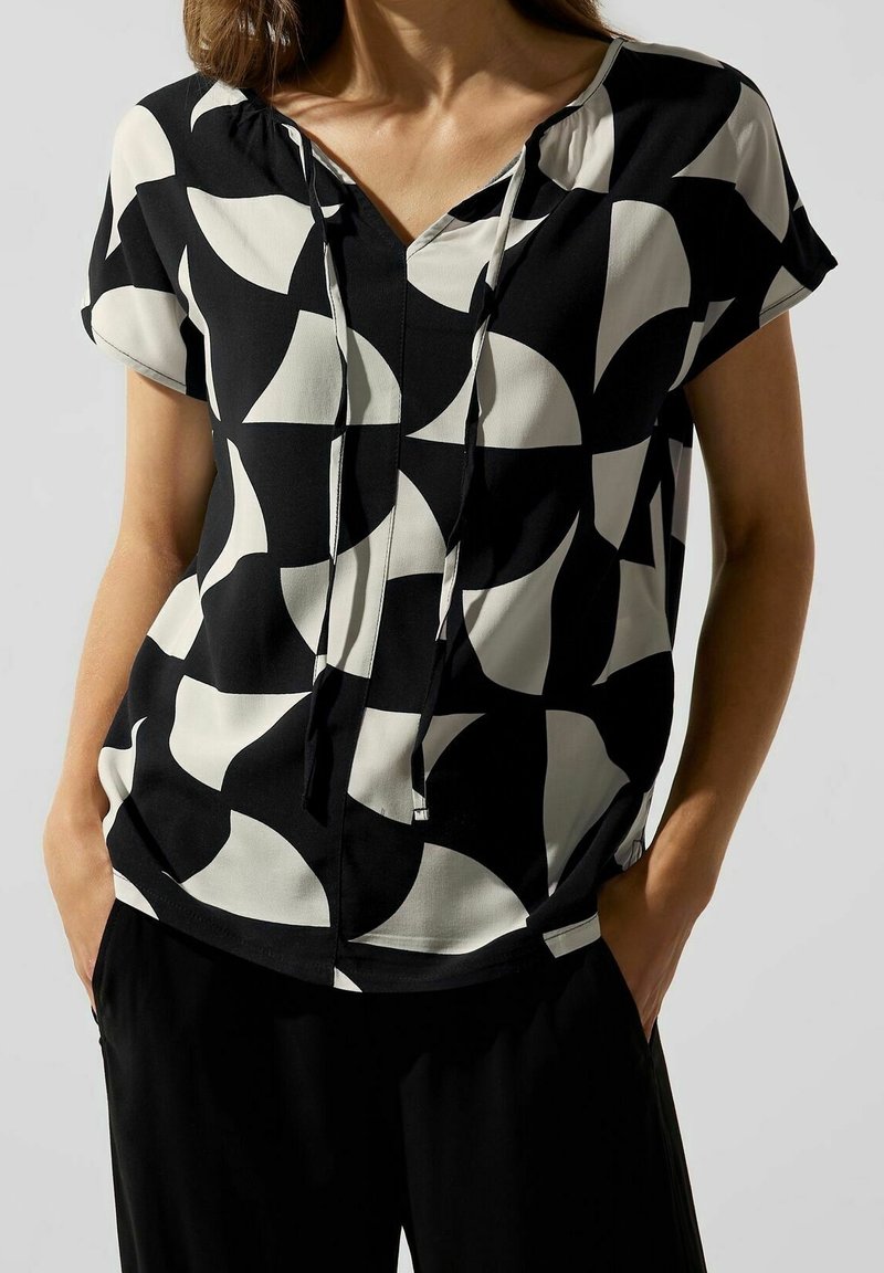 Haut en noir et blanc à motif, avec un décolleté en V, des manches courtes et des détails à cordon. Le tissu a une texture lisse et une coupe décontractée.