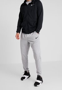 Felpa nera a zip, pantaloni grigi, entrambi con il logo Nike. La felpa presenta un cappuccio e tasche. Le scarpe da ginnastica nere completano l'outfit.