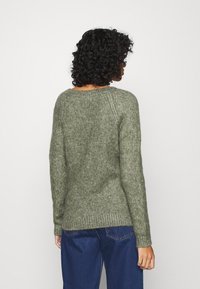 Grön stickad tröja med en texturerad yta, ribbad nederkant och raglanärmar, bärs med lösa blå jeans, sedd bakifrån.