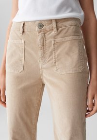 Persona che indossa pantaloni in velluto a coste beige con tasche anteriori a toppa e chiusura con bottone, abbinati a una camicia bianca infilata dentro.