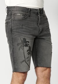 Shorts en denim gris foncé avec des imprimés de palmiers noirs et le texte « SUMMER VIBES BY KRSH » sur la cuisse gauche, portés par une personne en haut blanc.