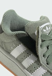 Zelené semišové tenisky s bílými koženými pruhy, texturovanými detaily a polstrovaným límcem. Obsahují logovou nášivku a robustní gumovou podrážku.