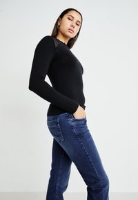 Schwarzes Langarm-Oberteil mit Spitzenakzenten an den Schultern, kombiniert mit blauen Jeans, im Seitenansicht mit den Händen in den Taschen.