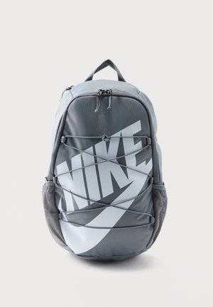 Szary plecak Nike z dużym białym logo, przednim elastycznym sznurkiem, siateczkowymi bocznymi kieszeniami i górnym uchwytem na białym tle.