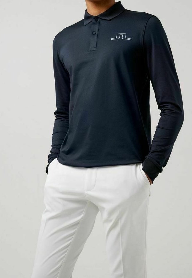 Dunkelblauses Langarm-Polohemd mit Kragen, versehen mit einem weißen Logo auf der Brust, kombiniert mit weißen Hosen aus glattem Stoff.