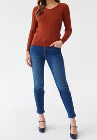 Femme portant un pull en maille torsadée couleur rouille, un jean bleu et des escarpins Mary Jane vernis bleu marine, debout avec une main dans la poche.