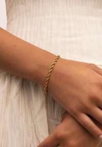 Pulsera de cadena de cuerda retorcida de oro llevada en la muñeca sobre piel clara, descansando contra un fondo de tela crema texturizada.