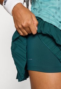 Teal sportieve rok met gelaagde plooien, met ClimaCool merk. Close-up van een hand die de stof aan de linkerkant aanpast.
