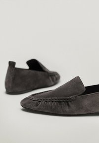 Mocassins en suède gris avec une semelle plate, dotés d'un bout arrondi et de coutures décoratives le long du bord supérieur. Modèle à enfiler, sans quincaillerie visible.