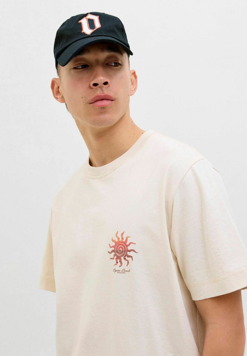T-shirt en coton beige avec un graphique de soleil rouge sur la poitrine gauche. Casquette noire avec un logo blanc bien visible. Design simple et décontracté.