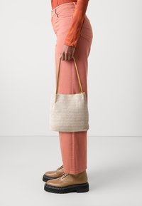 Osoba w koralowych spodniach i pomarańczowej bluzie z długim rękawem, trzymająca beżową plecioną torebkę z jasnobrązowymi ramiączkami, nosząca jasnobrązowe buty zapinane na sznurowadła z czarnymi podeszwami.