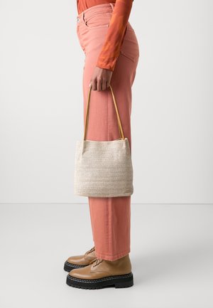 Persona con pantaloni corallo e maglia a maniche lunghe arancione, che tiene una borsa di paglia beige con manici color sabbia, indossa scarpe di corda beige con suola nera.