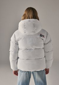 Giacca imbottita bianca con cappuccio ampio, con un logo sul retro e polsini elastici. Indossata sopra jeans azzurri.