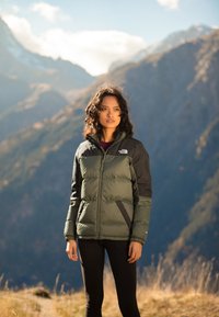 Steppjacke mit einem dunkelgrünen Körper und schwarzen Ärmeln, ausgestattet mit einem hohen Kragen, Reißverschluss und elastischen Ärmeln. Getragen über schwarzen Leggings.