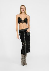 Bralette en dentelle noire avec des bords festonnés, associé à une jupe en dentelle noire avec des boutons dorés et des poches latérales, complété par des bottes en cuir laminé imprimé serpent.