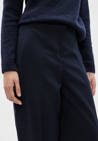 Χέρια δίπλα σε σκούρο μπλε παντελόνι καλοραμμένο και σε υφασμάτινη σκούρη μπλε μπλούζα, εστίαση στη μέση και στη λεκάνη με ορατό τατουάζ στον δεξί καρπό.