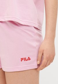 Shorts en coton rose avec un logo FILA rouge, présentant une texture lisse et une coupe décontractée. L'ourlet est légèrement roulé avec des coutures soignées.
