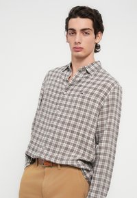 Πουκάμισο με μακριά μανίκια και κουμπιά, σε γκρι και καφέ καρό σχέδιο. Κατασκευασμένο από απαλό ύφασμα, με κλασικό γιακά και μικρή λεπτομέρεια με λογότυπο.